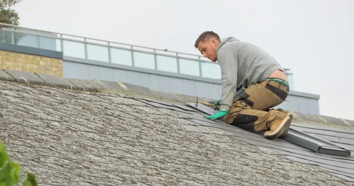 Roofer kneeling on a pitched shingle roof while inspecting and repairing roofing materials